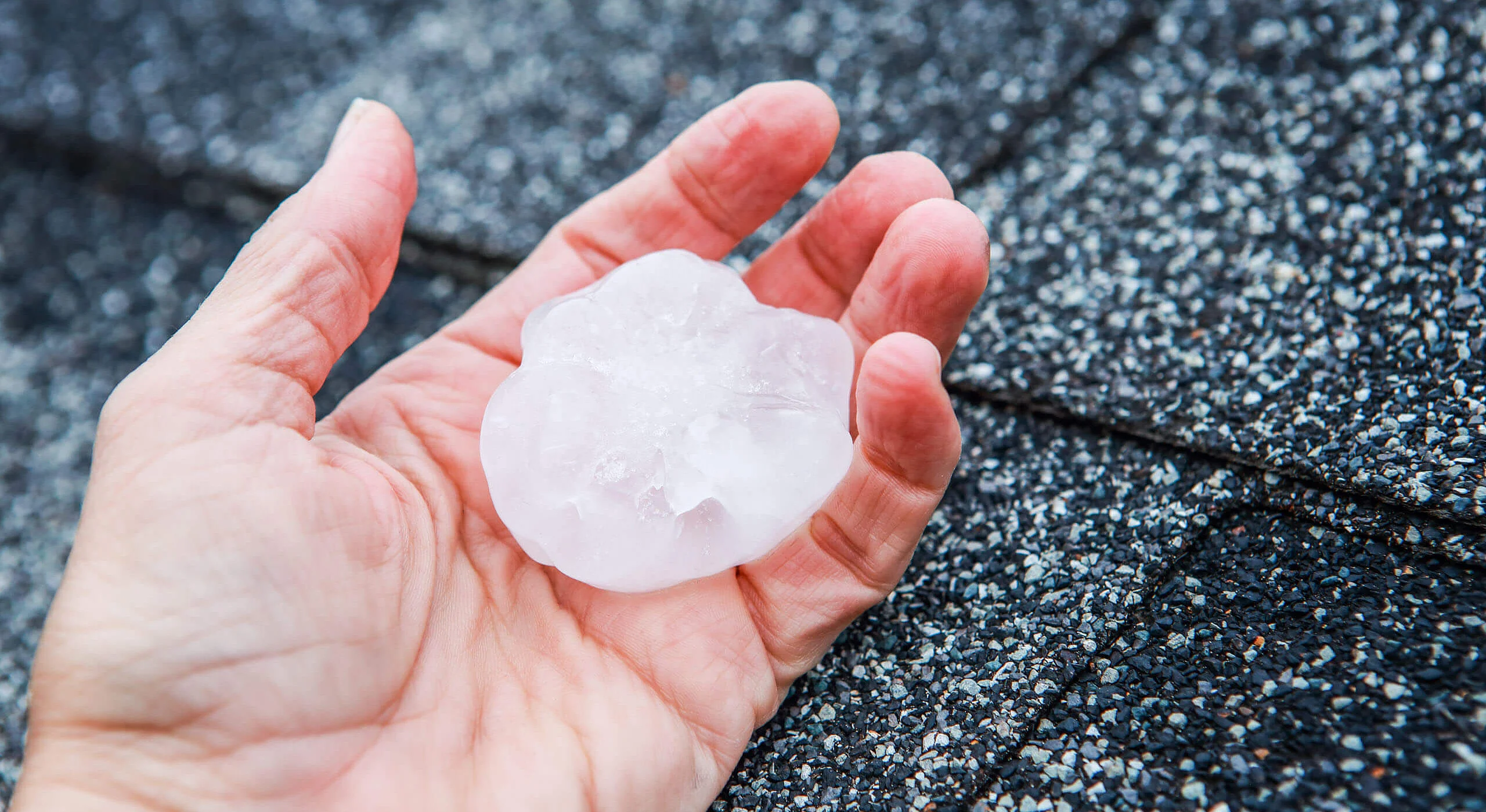 Großes Hagelkorn liegt in einer Hand, im Hintergrund ein Dach